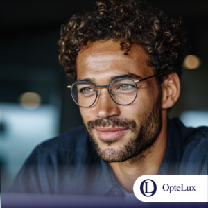 A man with curly hair and a beard wearing stylish round black-rimmed prescription glasses from OpteLux. He is looking thoughtfully to the side in a lifestyle setting. The OpteLux logo is visible in the bottom right corner